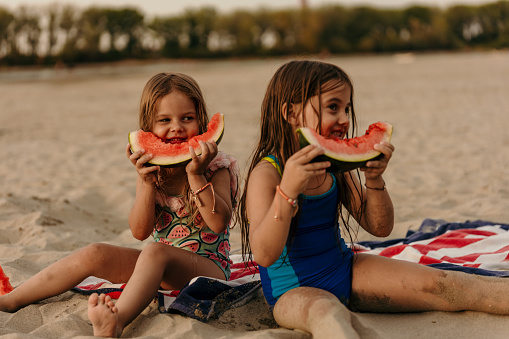 Two little girl children sitting wet on the sand, eating watermelon on the beach after a swimming, summertime refreshment.