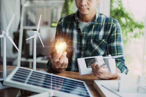 istock Close-up at a light bulb, An engineer sits holding a light bulb and model of wind turbine. with solar panels and model house on the table. To design use of renewable energy with wind and solar energy. 1419789912