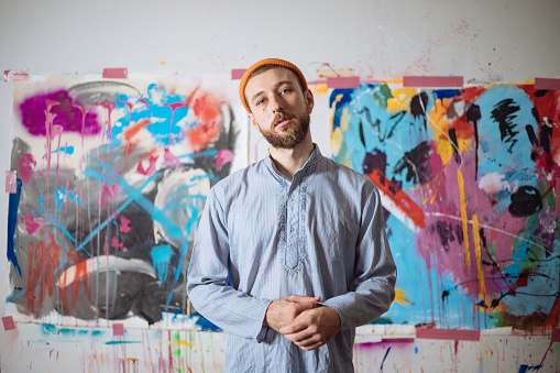 A handsome Israeli artist stands in front of his abstract paintings displayed on a wall in his studio behind him.