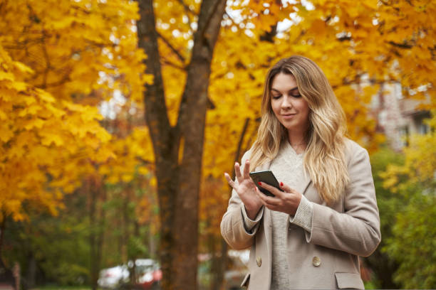 una hermosa mujer en un parque de otoño con un abrigo ligero mira algo en un teléfono inteligente. árboles amarillos y hojas caídas. - dropped call fotografías e imágenes de stock