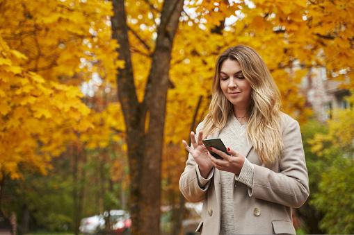 a beautiful woman in an autumn park in a light coat looks at something in a smartphone. yellow trees and fallen leaves. High quality photo