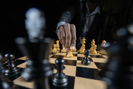 Close-up of man's hand moving pawn chess piece on chessboard against black background.