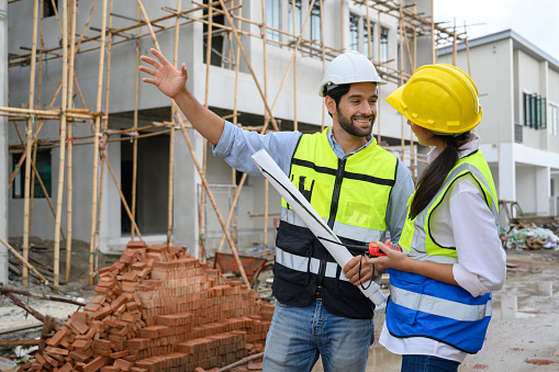 Young attractive construction man and woman in vests with helmets working on the under-construction building site. Home building project. Engineer foreman discusses with a coworker at workplace
