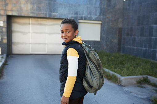 Rare view of cute happy african american kid of 12 returning home from school wearing puffer vest and backpack on shoulders standing outside near underground parking of his neighbor