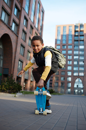 Cute african american kid going to school by his blue longboard, wearing backpack on his shoulders, standing on skateboard in ready position, looking at camera in his neighborhood