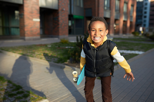 Black boy kid with skateboard in hands roaring at camera showing teeth on city street background, making face, grimacing, expressing his attitude about going to school, carrying backpack on shoulders