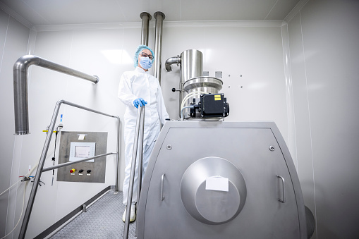 Pharmaceutical technician in a protective clothing seen standing in a laboratory by one of the special machines during a manufacturing process.