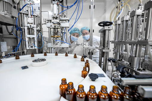 Small brown bottles seen perfectly arranged in a laboratory machine during manufacturing in a pharmaceutical factory while two female employees in a protective clothing are controlling the process.
