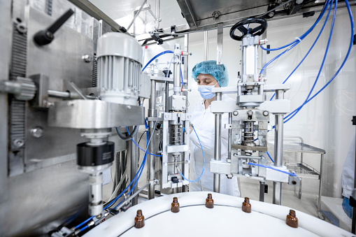 One employee in pharmaceutical industry wearing protective gloves, mask, cap and white suit seen standing by the machine that is the part of the medicaments production during the working hours in a pharmaceutical manufacturing.