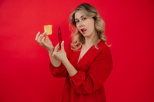Playful blonde woman with sticking tongue hold hot spicy pepper and salty potato chips on red isolated background. Fast food snacks choice and body positive. Spice hot flavor for stomach. Copy space