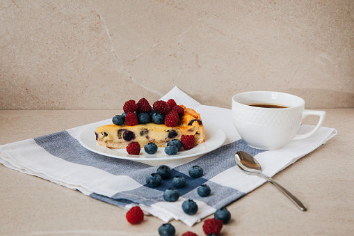 A piece of blueberry cheesecake on a white plate with fresh raspberries and blueberries and a cup of coffee on a stone table. Front view