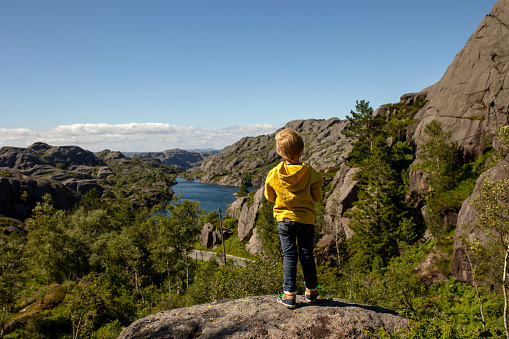 A young boy hiker is standing on the cliff admiring the view over Oeschinensee Lake near Kandersteg in Berner Oberland region in Switzerland, where he is hiking with his family. The boy is wearing a sun hat.