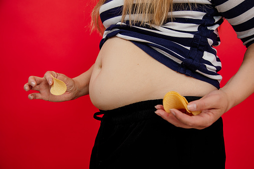 Mujer adiposa recortada con vientre desnudo en ropa a rayas sosteniendo, comiendo papas fritas. Alimentos chatarra poco saludables que engordan. photo