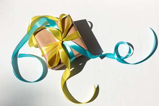 The gift is wrapped in kraft paper with yellow and blue ribbon with shadow on a white background. Top view