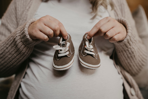 Expectant mother standing in studio holding up cute baby shoes, walking in your shoes. High quality photo