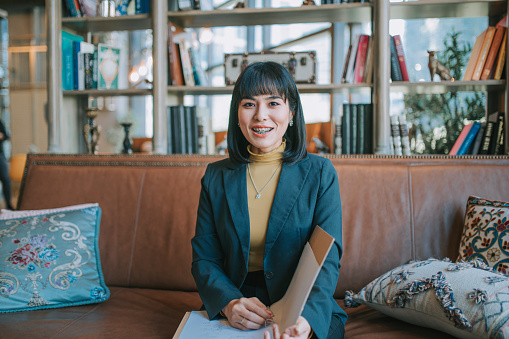 Asian Malay Woman with dental braces looking at camera smiling sitting on sofa in business lounge holding file folder