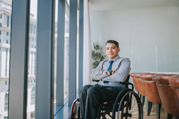 confianza persona discapacitada hombre de negocios asiático sentado en los brazos cruzados de la silla de ruedas mirando a la cámara sonriendo en la sala de conferencias - physical impairment smiling front view looking at camera fotografías e imágenes de stock