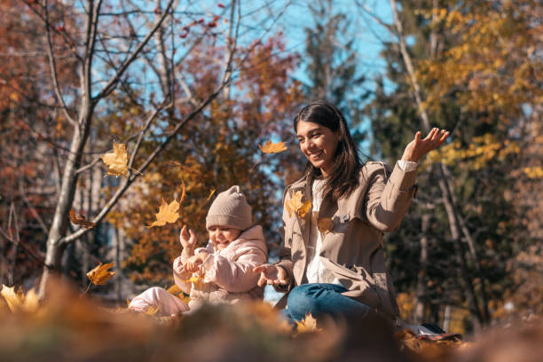 Happy dark-haired young mother and daughter having fun in the autumn park, throwing yellow leaves on a sunny day.Family and autumn concept stock photo