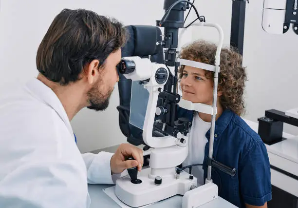 Cute boy while eye exam with binocular slit-lamp at ophthalmology clinic with experienced optometrist. Vision correction in children