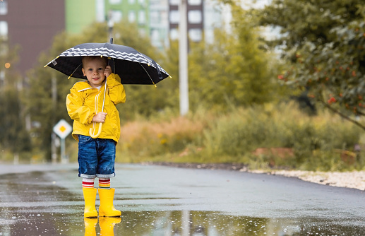 A cute little boy 3 years old jumps through puddles and laughs merrily. A picture of summer and autumn holidays. A child plays outside in the rain in a yellow raincoat and boots .