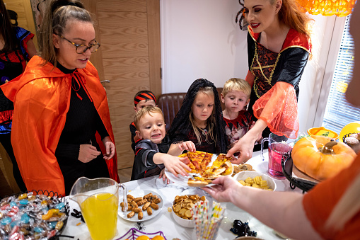 Friends and family stand around a table filled with Halloween-related food and drink. People reach out for pizza.