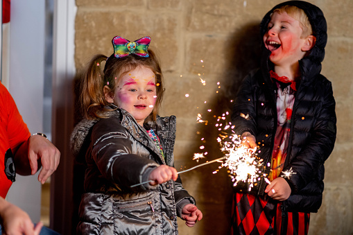 A young girl holding a sparkler excitably wearing a warm jacket in the garden. A young boy is standing next to her laughing also wearing a warm jacket.