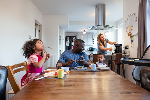 Family having breakfast in the morning together in the North East of England. The father sitting with his young children at the table. They are sharing fruit and toast while the woman is busy in the kitchen behind them. The little girl is playing with a spoon in her mouth. The man is living with a disability after having polio.