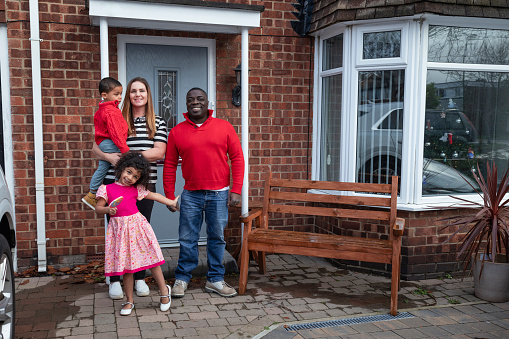 Family with two children standing outside of their home in the North East of England. They are smiling at the camera and it is around Christmas time, as they have a Christmas tree in their window.