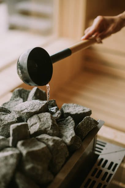 main d’une jeune femme versant de l’eau sur des roches chaudes dans le sauna - sauna photos et images de collection