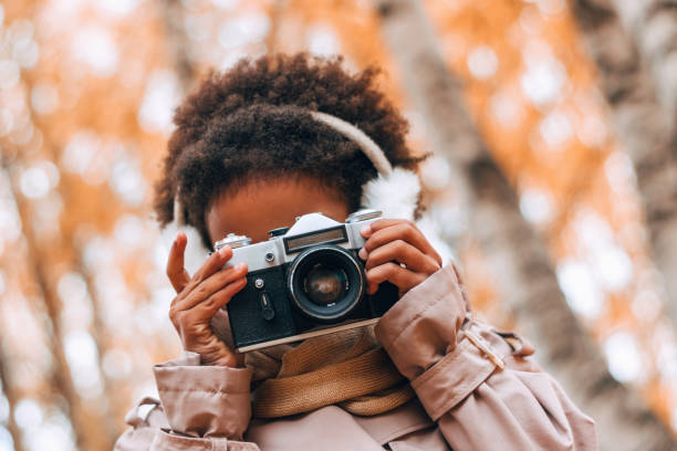 모피 헤드폰을 입은 귀여운 아프리카 계 미국인 소녀가 가을 공원에서 카메라로 사진을 찍습니다. 다양성, 가을 개념 - photographer enjoyment elegance old fashioned 뉴스 사진 이미지