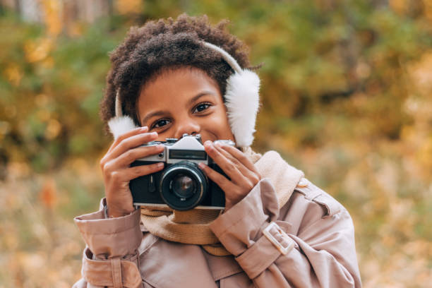 Cute happy African-American girl in fur headphones takes pictures with a camera in an autumn park.Diversity,autumn concept