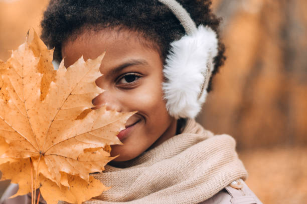 Portrait of a cute African-American girl in fur headphones, covering her face with a yellow maple leaf in an autumn park.Diversity,autumn concept stock photo