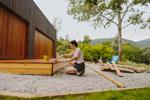 Photo of a young woman painting a wooden deck in front of the house