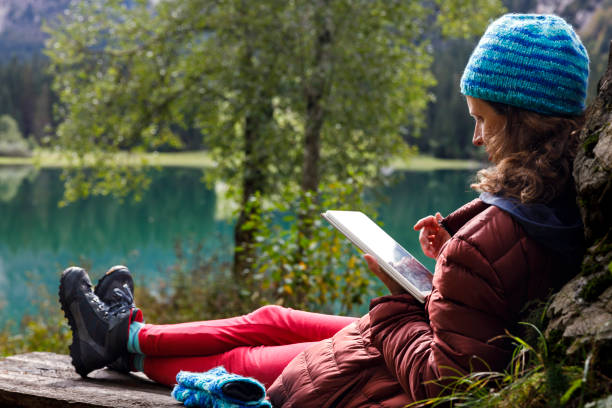 wanita dewasa tengah belajar melalui tablet digital di luar ruangan di bangku dekat danau alpine yang indah - wilderness studies potret stok, foto, & gambar bebas royalti