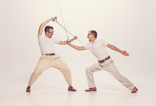 Portrait of two men in a suit fighting with swords isolated over grey studio background. Expressing emotions. Concept of sport, hobby, emotions, active lifestyle, retro fashion. Copy space for ad