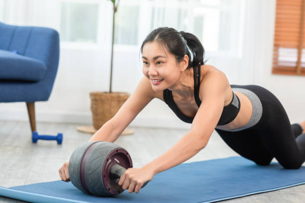 アスリートアジアの若い女性は自宅で運動マットにabローラーで運動します。強い筋肉を構築するためにabホイールローラーのスポーツウェアワークアウト運動バランスボディで健康な女性。 - 腹筋ローラー ストックフォトと画像