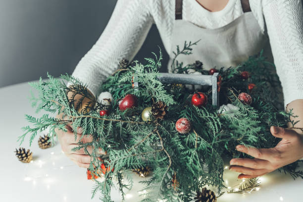 istock Florist woman makes Christmas composition for table decoration from thuja, spruce branches in wooden box. Using of floral foam, cones, snow apples, balls, cotton flowers,garland. New Year celebration 1419889016