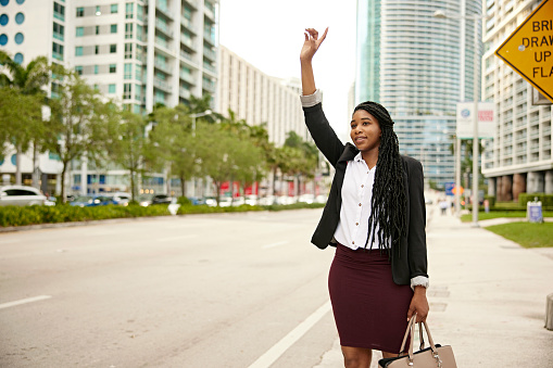 Front view of mid 20s businesswoman wearing corporate attire, standing curbside and gesturing, with business district in background.
