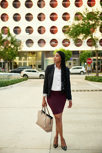 Full length front view of young Black professional in corporate attire holding handbag, standing with legs crossed, and looking away from camera.