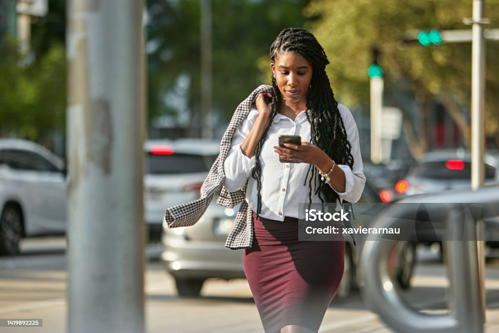 Businesswoman checking smart phone while walking downtown Front view of longhaired Black woman in mid 20s approaching camera with jacket on shoulder and looking at portable device. African-American Ethnicity Stock Photo