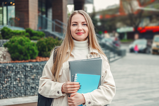 Education concept. Happy student girl holds folders notebooks books in hands smiles look at camera against the background of a modern university building