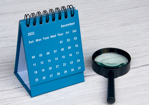 December 2022 blue color desk calendar on wooden desk with copy space and magnifying glass.