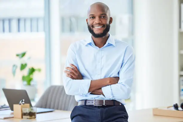 Photo of Confident business man standing with arms crossed in an office, looking proud and happy alone at work. Portrait of a smiling, cheerful and professional African male boss working in corporate