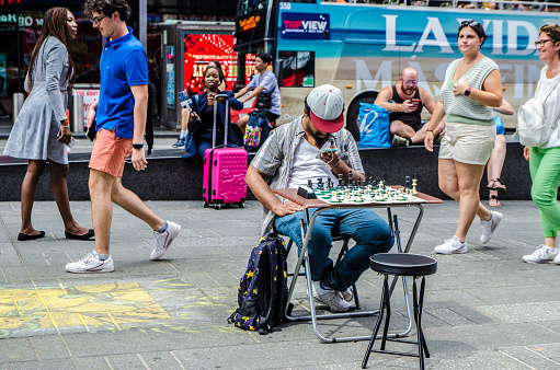 Black man playing chess alone in the middle of the walkway in New York during summer day.\nIs he waiting for someone to sit with him to play ?