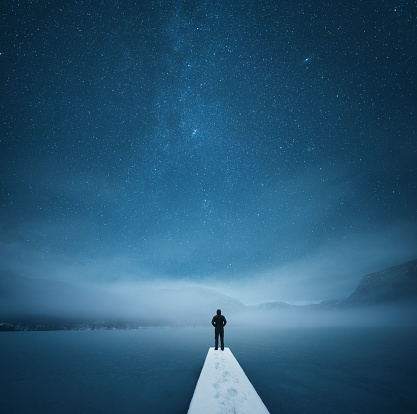 Peaceful winter night by the frozen lake. Man is standing on the pier and watching stars.