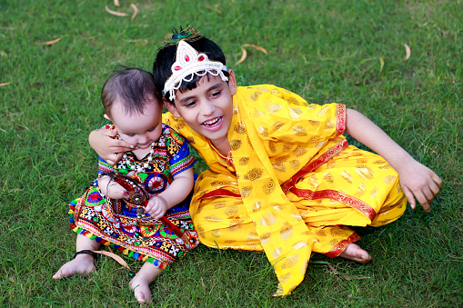 Cute little siblings of Indian ethnicity sitting portrait  in the park wearing fancy dress during shri krishna  janmashtami festival outdoor in nature during spring time.