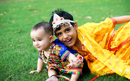 Cute little siblings of Indian ethnicity sitting portrait  in the park wearing fancy dress during shri krishna  janmashtami festival outdoor in nature during spring time.