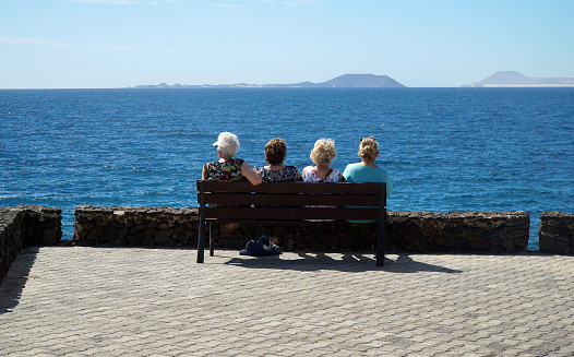 Playa Blanca, Lanzarote, Spain - March 27, 2022: Four older ladies sitting on bench   looking at view across sea to  Fuerteventura