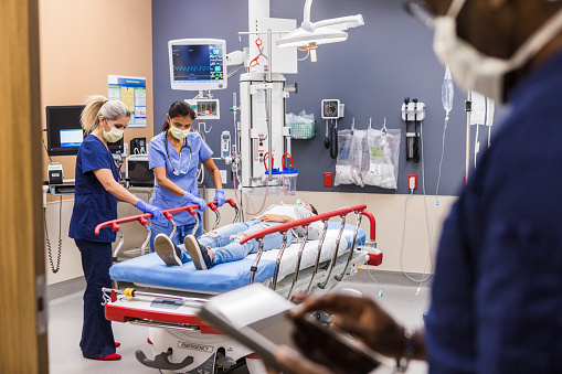 An unrecognizable male ER doctor in foreground uses a digital tablet to access the test results as two female nurses raise the side rails of a gurney holding a young adult female patient.