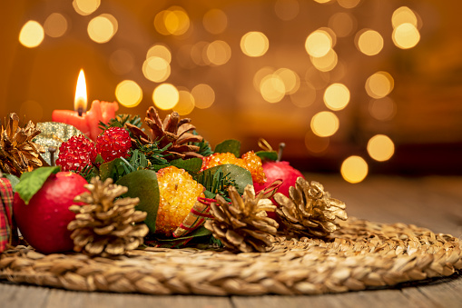 CHRISTMAS TABLE CENTERPIECE WITH RED CANDLE, FRUITS AND PINE CONES ON CATTAIL STRAW ROUND. BOKEH LIGHTS BACKGROUND. SELECTED FOCUS.